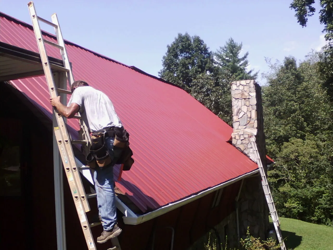 Roofer on a ladder beside a red metal roof and stone chimney — Soffit & Fascia Repair by Roofing Brothers in Salkum